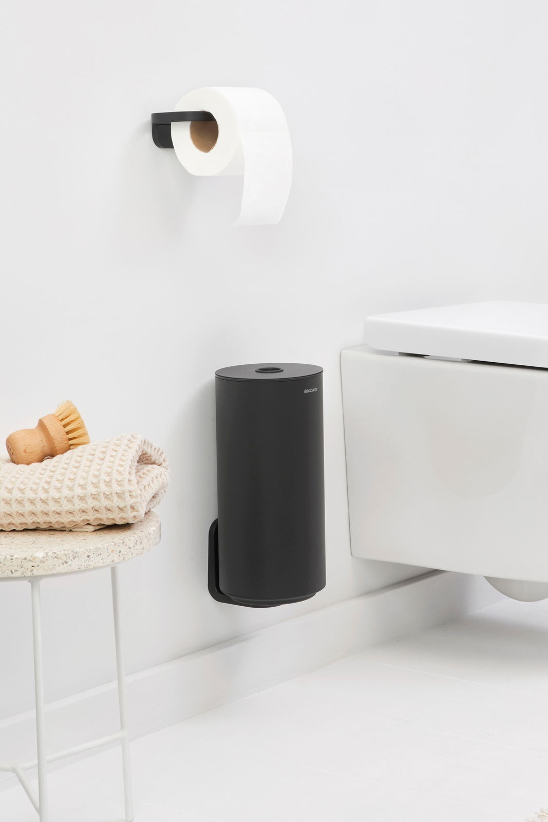 Schwarzer Toilettenrollenspender an der Wand montiert, neben einer weißen Toilette, mit einem modernen und minimalistischen Design. Perspektive von der Seite.