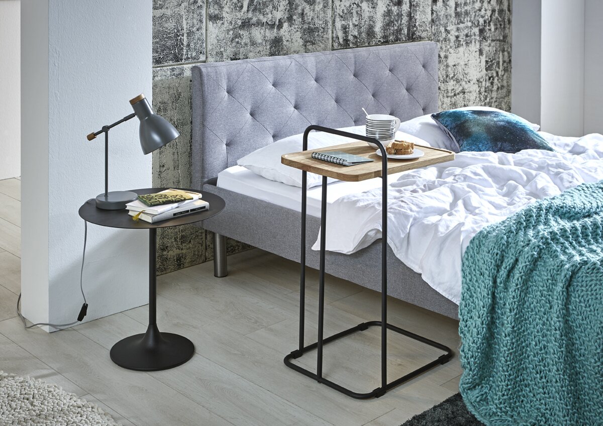 Moderner Beistelltisch/Serviertisch mit schwarzem Metallgestell und Holztablett als Ablage neben einem grauen Polsterbett, seitliche Perspektive im Schlafzimmer.