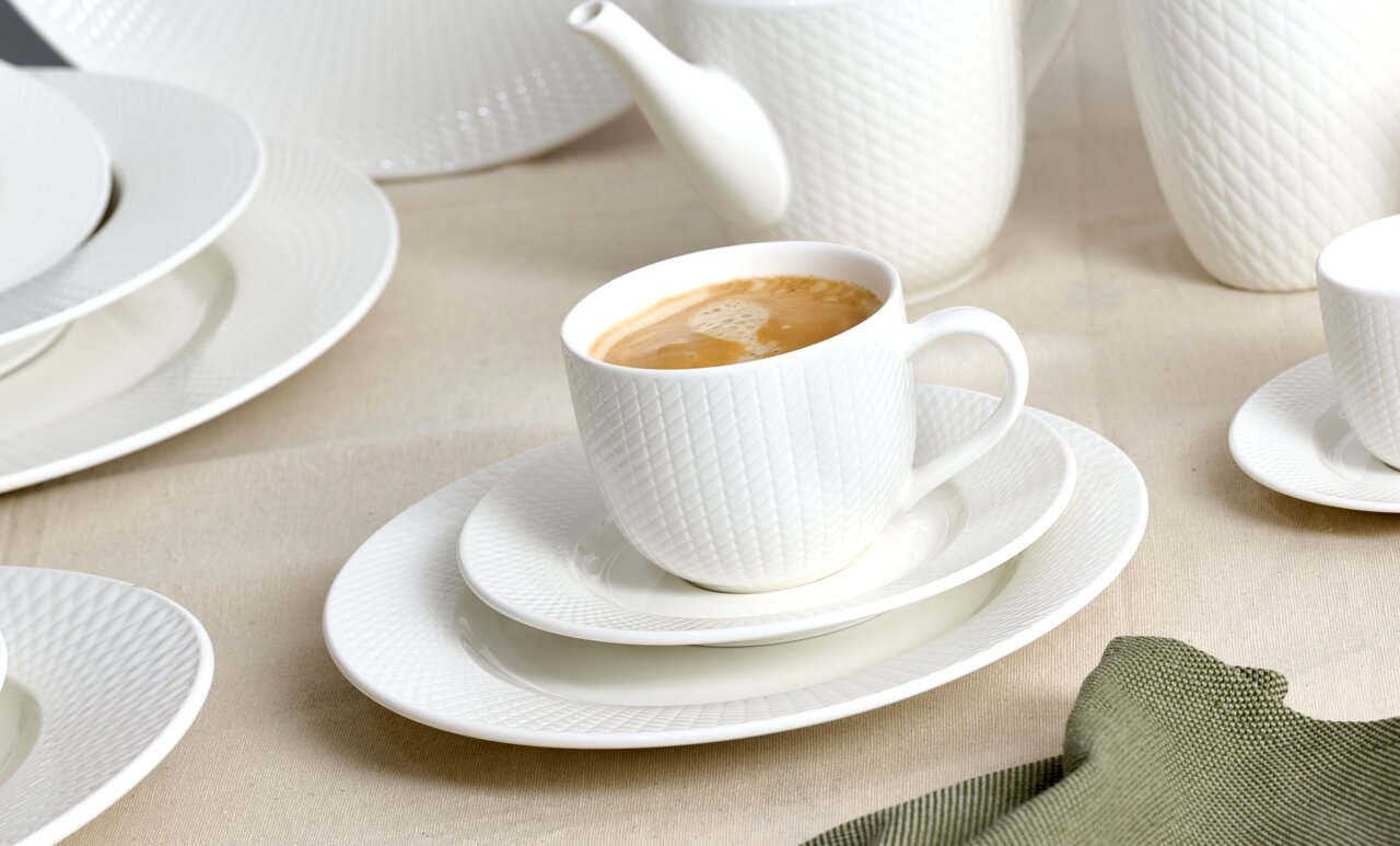 Weiße Kaffeeobertasse mit Untertasse, gefüllt mit Kaffee, auf einem Tisch mit weiteren Geschirrteilen, seitliche Perspektive