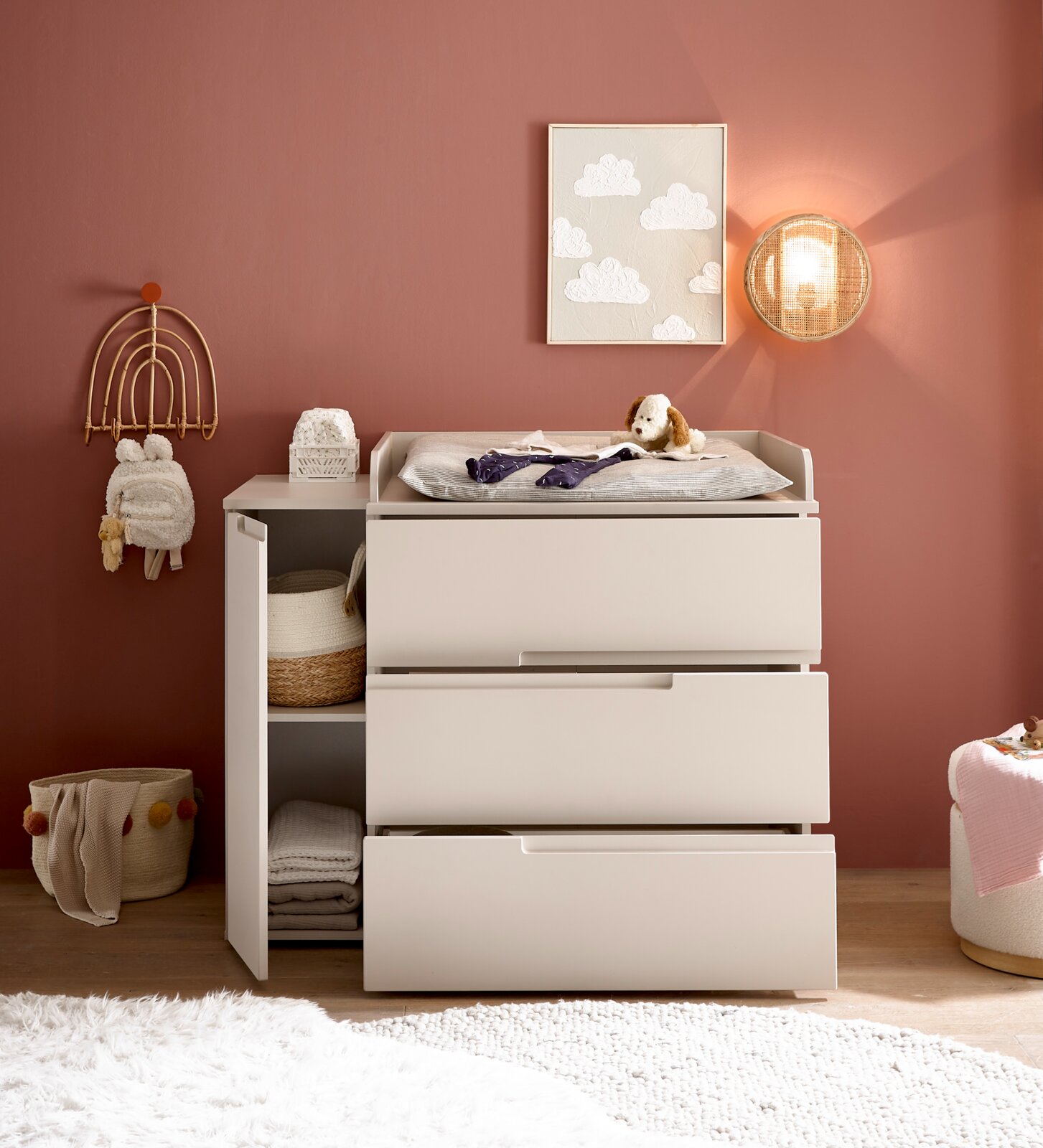 Weiße Wickelkommode mit drei Schubladen und offenem Regal, oben Wickelauflage mit Stofftier; Frontansicht in einem Kinderzimmer mit rosafarbener Wand, Wandbild und runder Wandleuchte.