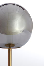 Nahaufnahme einer Tischleuchte mit kugelförmigem, rauchgrauem Glasschirm und einem goldenen Ständer, seitliche Perspektive.