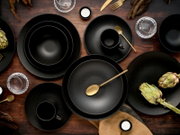 Schwarzes, mattes Geschirrset mit Tellern, Schalen und Tassen, stilvoll arrangiert mit goldenem Besteck, Gläsern und Artischocken auf einem dunklen Holztisch, aus der Vogelperspektive.