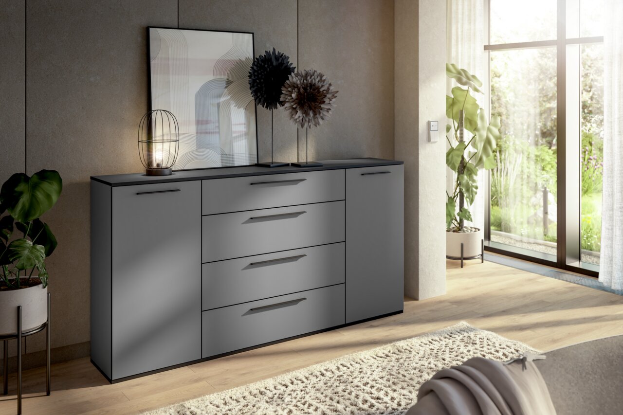 Modernes graues Sideboard mit Schubladen und Türen, dekoriert mit einer Lampe und Pflanzen, in einem hellen Raum mit Blick auf den Garten. Perspektive von vorne links.