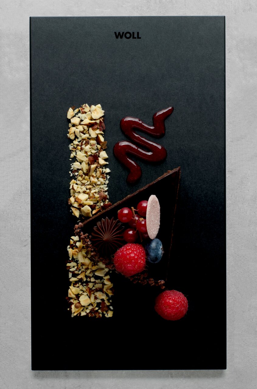 Vogelperspektive auf ein elegantes Frühstücksset mit einer schwarzen Platte, dekoriert mit einem Stück Schokoladenkuchen, Himbeeren, Johannisbeeren, Blaubeeren, gehackten Nüssen und einer Spur von roter Soße.