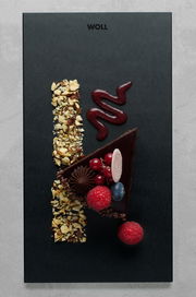 Vogelperspektive auf ein elegantes Frühstücksset mit einer schwarzen Platte, dekoriert mit einem Stück Schokoladenkuchen, Himbeeren, Johannisbeeren, Blaubeeren, gehackten Nüssen und einer Spur von roter Soße.