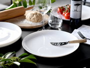 Pizza-Messer mit Holzgriff und Klinge mit „PIZZA“-Ausschnitt auf weißem Teller, schräg von oben auf gedecktem Tisch mit Teig, Tomaten, Weinglas und Flasche