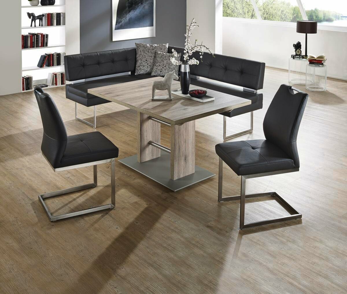 Moderner Esstisch mit Holzoptik und Metallgestell, umgeben von schwarzen Stühlen und einer Eckbank, in einem hellen Raum mit großen Fenstern, seitliche Perspektive.