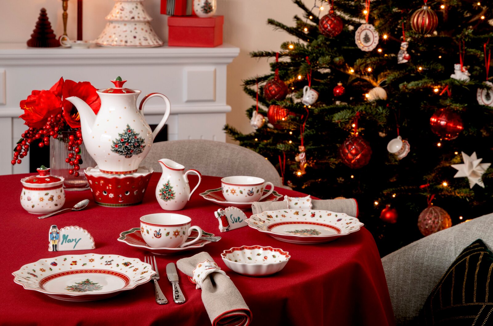 Festlich gedeckter Weihnachtstisch mit weiß-rotem Porzellanservice, Kanne, Tassen und Tellern mit Weihnachtsmotiven, aus schräger Vorderansicht vor einem beleuchteten Christbaum.