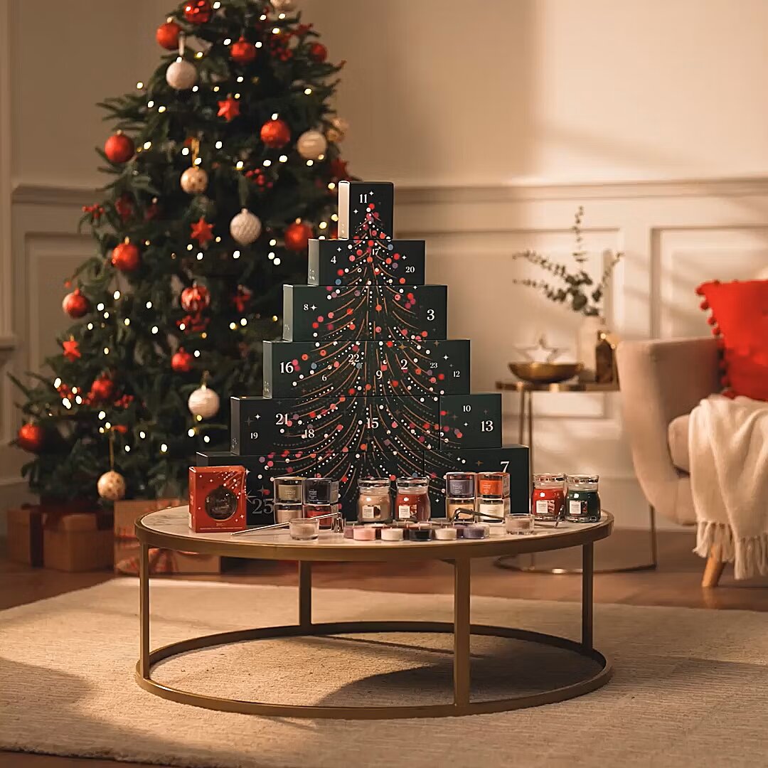 Frontalansicht eines dekorativen Adventskalenders in Weihnachtsbaumform mit 25 nummerierten Fächern und kleinen Duftkerzen auf einem Couchtisch vor einem festlich geschmückten Weihnachtsbaum.