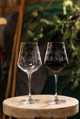 Zwei Rotweingläser mit winterlichen Gravuren, eines gefüllt mit Rotwein, auf einem rustikalen Holztisch, umgeben von natürlicher Dekoration. Seitliche Perspektive.