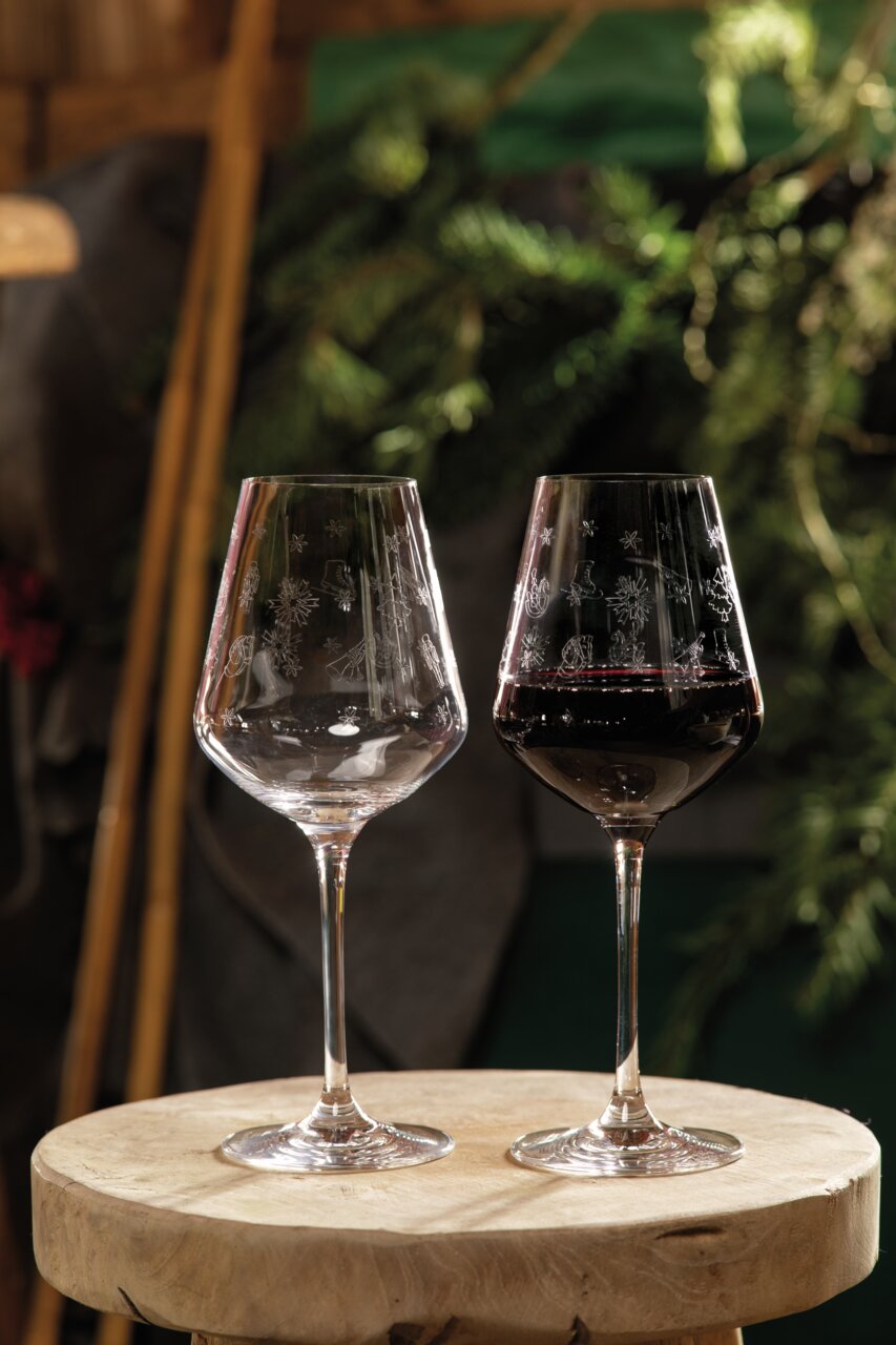 Zwei Rotweingläser mit winterlichen Gravuren, eines gefüllt mit Rotwein, auf einem rustikalen Holztisch, umgeben von natürlicher Dekoration. Seitliche Perspektive.