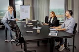 Moderne Büroumgebung mit drei Personen, die an einem großen Tisch arbeiten, auf ergonomischen Drehstühlen sitzend. Perspektive von der Seite.