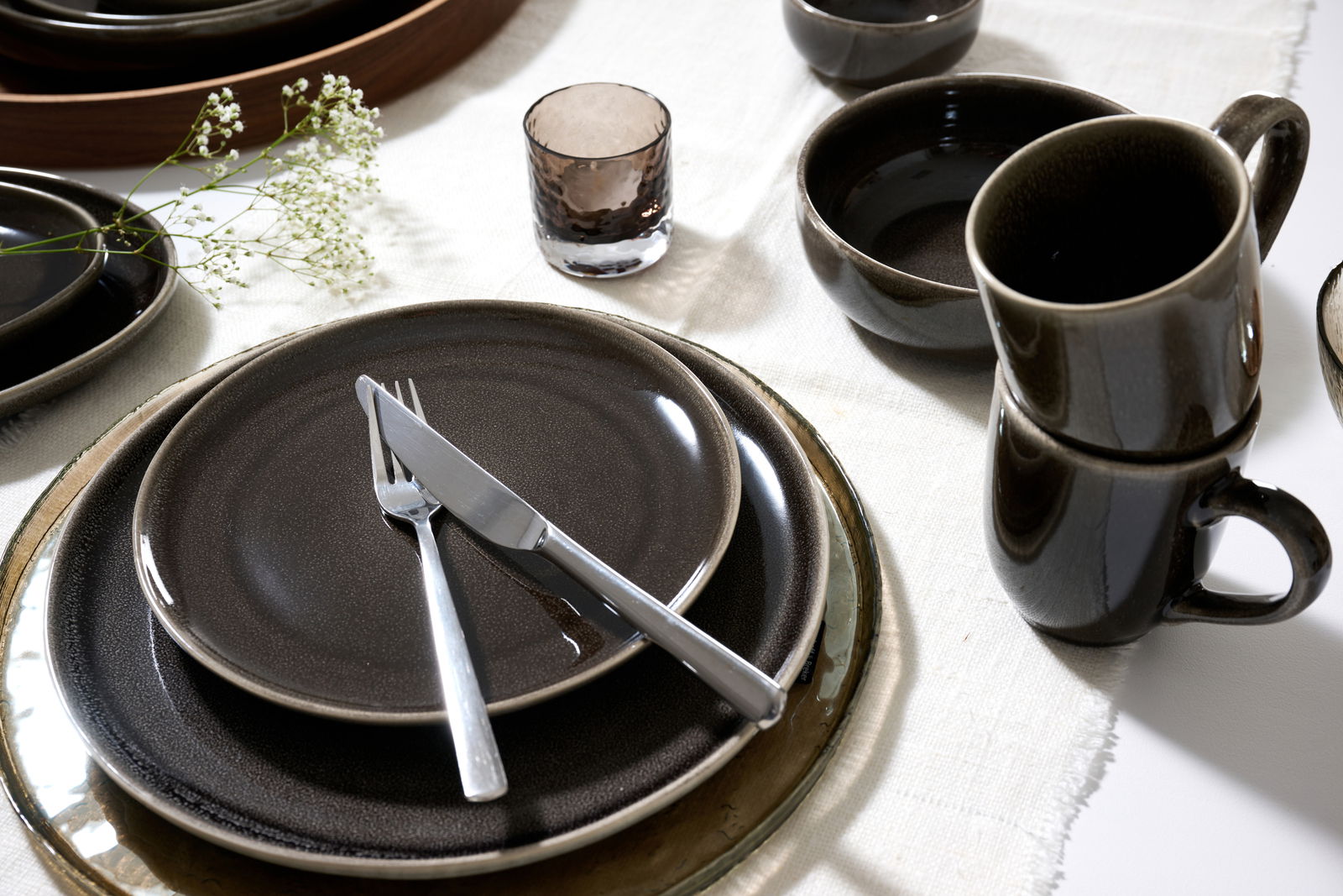 Elegantes Geschirrset in dunklen Tönen mit Tellern, Tassen und Schalen, von oben betrachtet, auf einem weißen Tischtuch arrangiert.