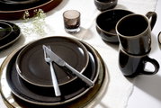 Elegantes Geschirrset in dunklen Tönen mit Tellern, Tassen und Schalen, von oben betrachtet, auf einem weißen Tischtuch arrangiert.