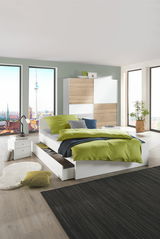 Modernes Schlafzimmer mit einem weißen Bett mit grüner Bettwäsche, Schubladen an der Seite und einem großen Fenster mit Stadtblick. Perspektive: seitlich von vorne.