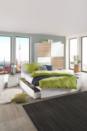 Modernes Schlafzimmer mit einem weißen Bett mit grüner Bettwäsche, Schubladen an der Seite und einem großen Fenster mit Stadtblick. Perspektive: seitlich von vorne.