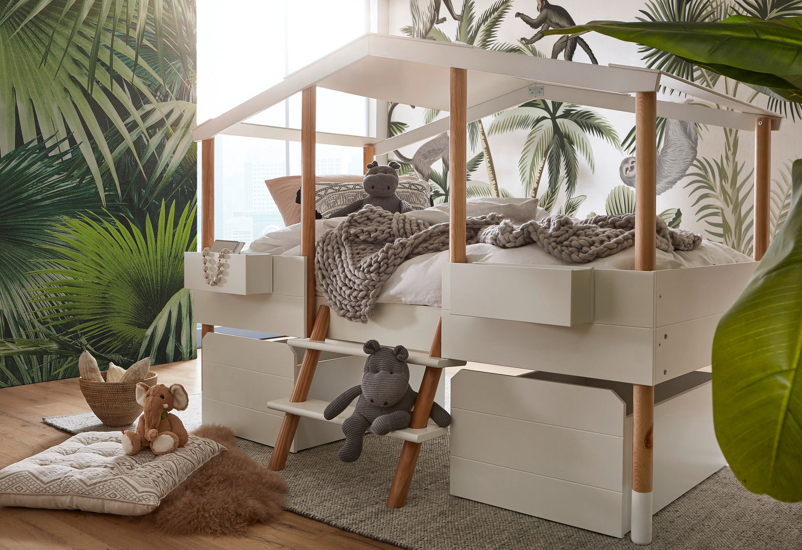 Hausbett mit Holzpfosten und integriertem Stauraum, dekoriert mit Kuscheltieren und Decken, in einem Kinderzimmer mit Dschungelmotiv-Tapete. Perspektive von schräg vorne.