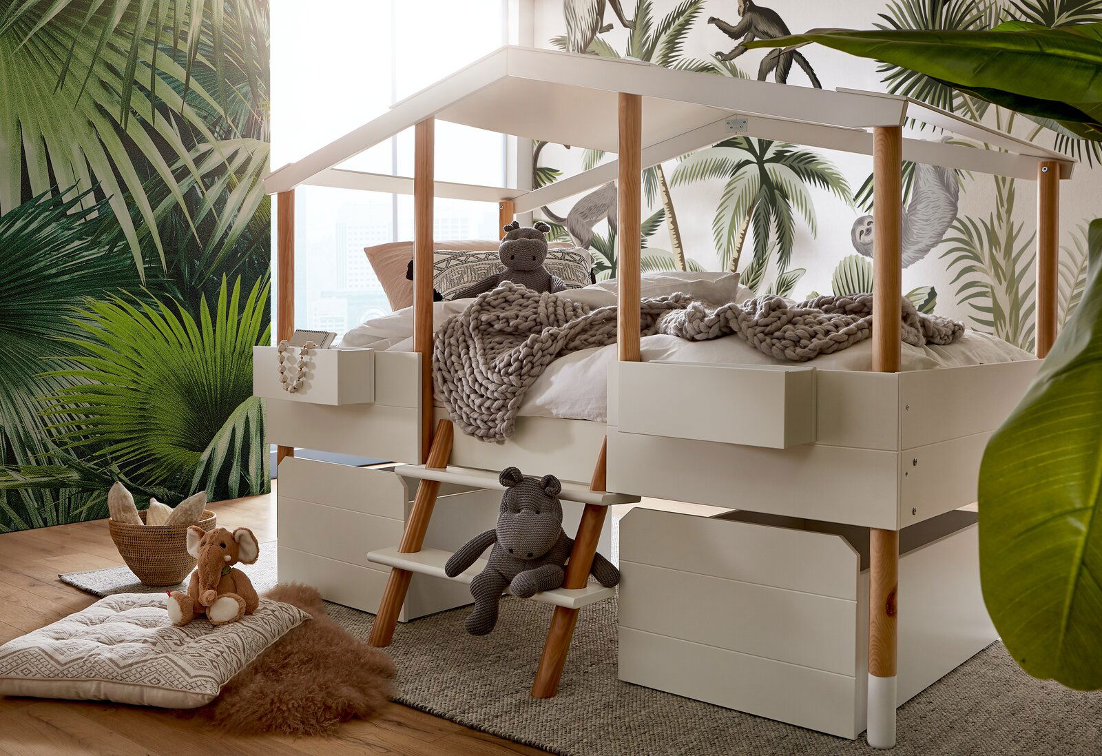 Hausbett mit Holzpfosten und integriertem Stauraum, dekoriert mit Kuscheltieren und Decken, in einem Kinderzimmer mit Dschungelmotiv-Tapete. Perspektive von schräg vorne.