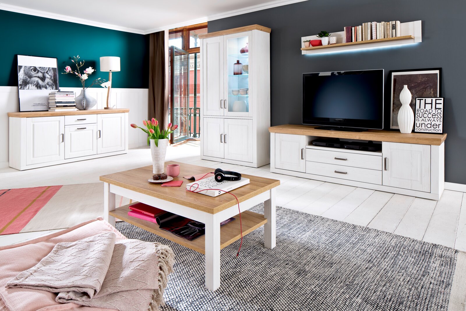 Helles Wohnzimmer-Set im Landhausstil mit weißer Anrichte, Vitrine, TV-Lowboard und Couchtisch in Weiß mit Holzoptik-Platten, frontal in leichter Weitwinkelperspektive fotografiert.