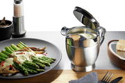 Thermo-Sauciere SERVIRO aus Edelstahl mit geöffnetem Deckel, gefüllt mit Sauce, auf einem Tisch neben einem Teller mit Spargel und einer Käsereibe. Perspektive von vorne.