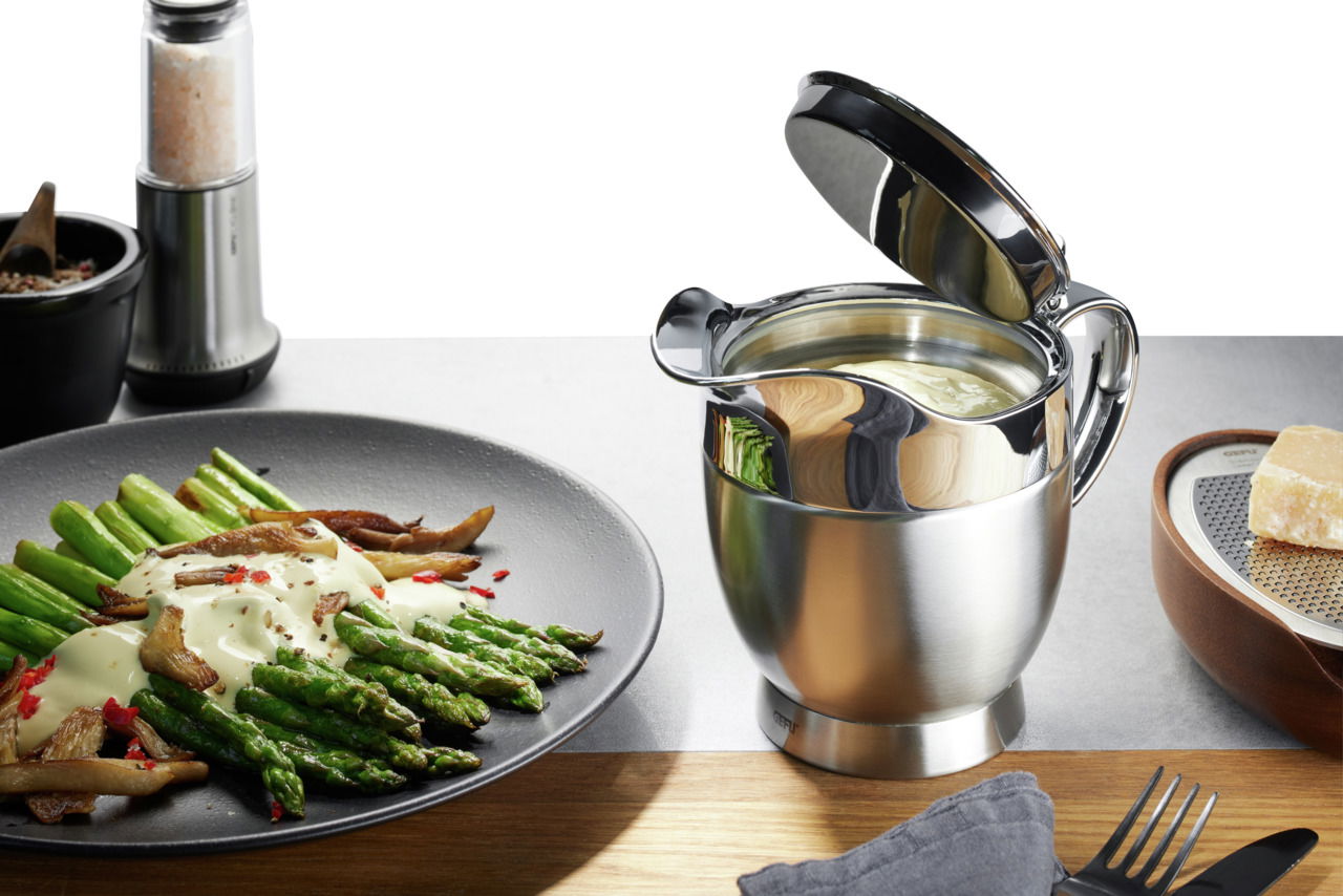 Thermo-Sauciere SERVIRO aus Edelstahl mit geöffnetem Deckel, gefüllt mit Sauce, auf einem Tisch neben einem Teller mit Spargel und einer Käsereibe. Perspektive von vorne.