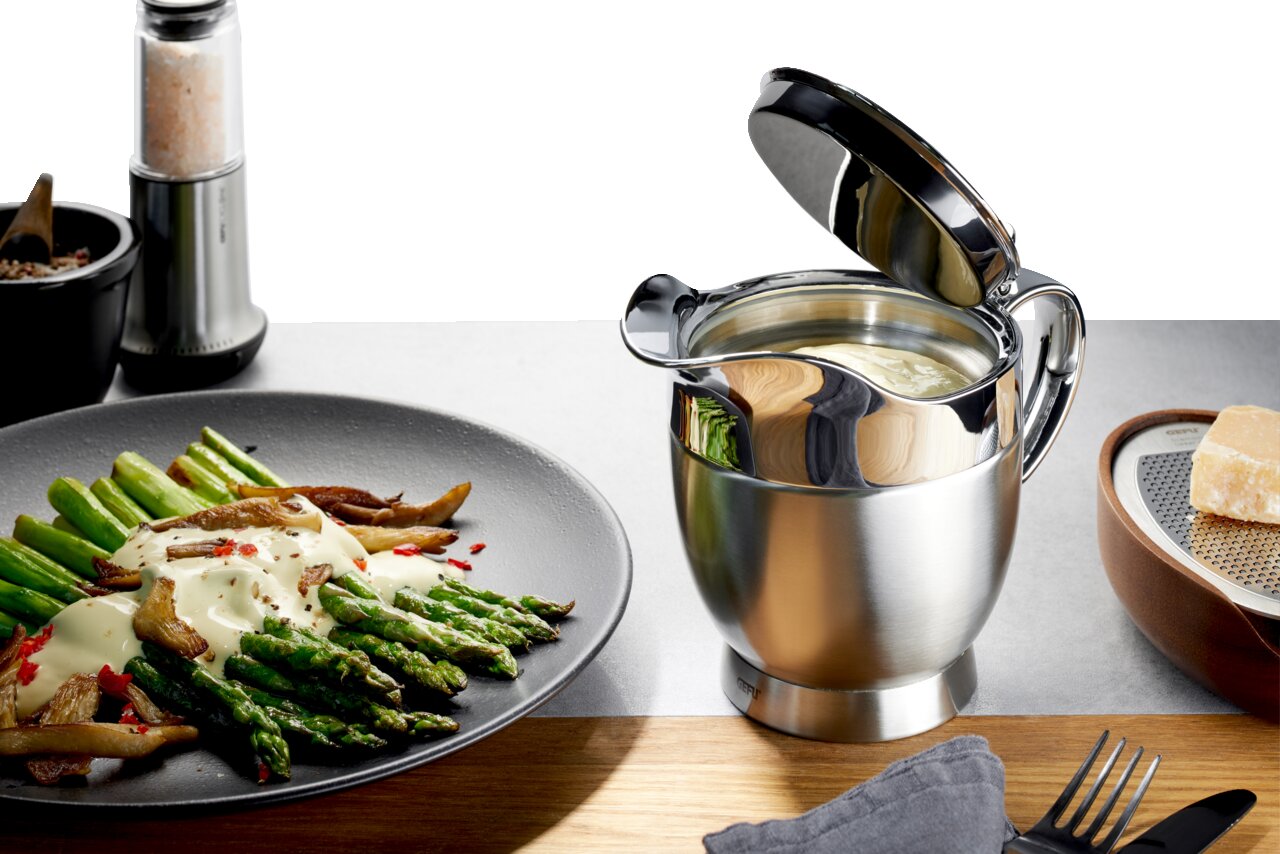 Thermo-Sauciere SERVIRO aus Edelstahl mit geöffnetem Deckel, gefüllt mit Sauce, auf einem Tisch neben einem Teller mit Spargel und einer Käsereibe. Perspektive von vorne.