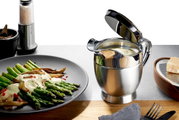 Thermo-Sauciere SERVIRO aus Edelstahl mit geöffnetem Deckel, gefüllt mit Sauce, auf einem Tisch neben einem Teller mit Spargel und einer Käsereibe. Perspektive von vorne.