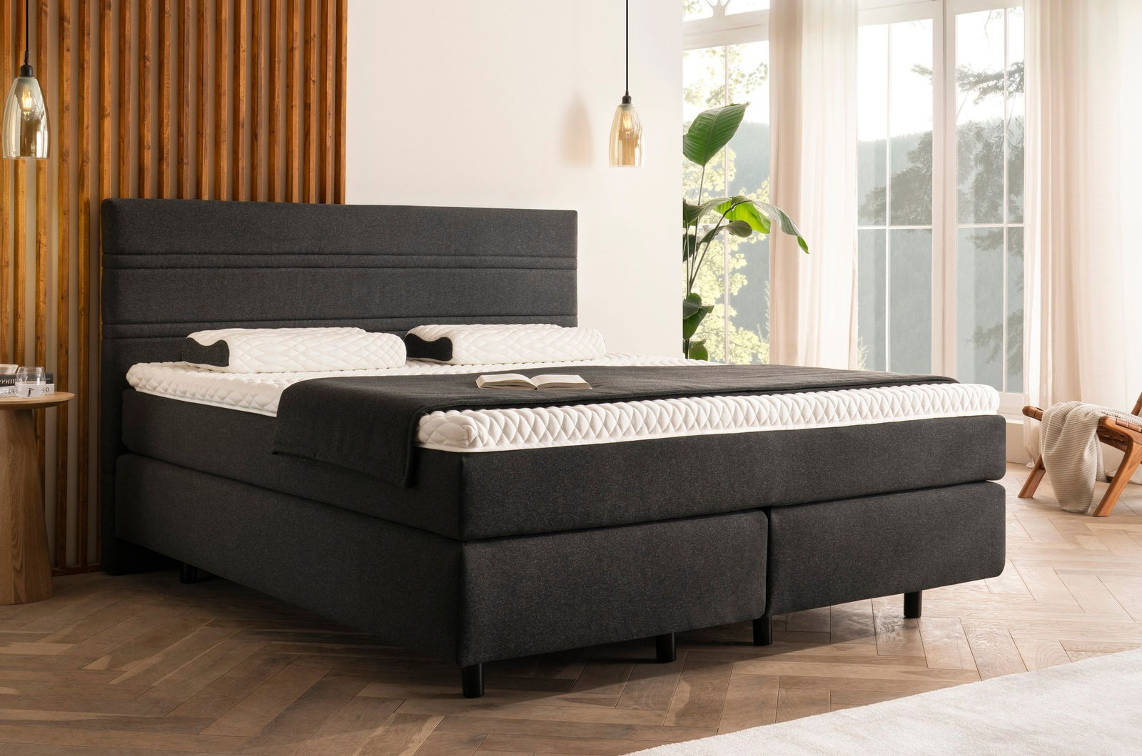 Boxspringbett in einem modernen Schlafzimmer, seitliche Perspektive, mit dunklem Stoffbezug und heller Bettwäsche, umgeben von Holzverkleidung und großen Fenstern.
