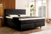 Boxspringbett in einem modernen Schlafzimmer, seitliche Perspektive, mit dunklem Stoffbezug und heller Bettwäsche, umgeben von Holzverkleidung und großen Fenstern.