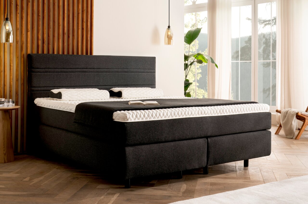 Boxspringbett in einem modernen Schlafzimmer, seitliche Perspektive, mit dunklem Stoffbezug und heller Bettwäsche, umgeben von Holzverkleidung und großen Fenstern.