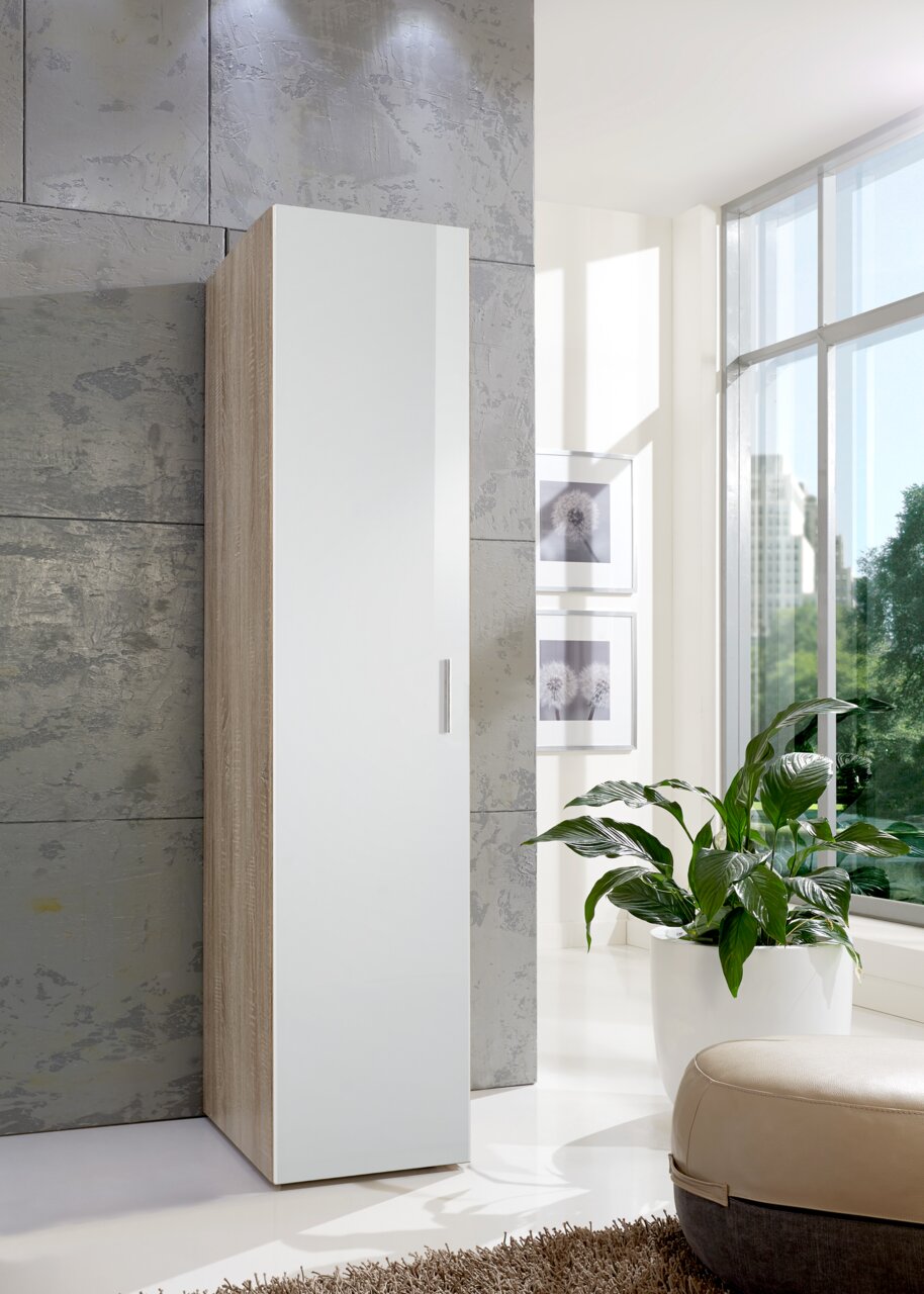 Moderner, schmaler Kleiderschrank in Holzoptik mit weißer Tür, seitliche Perspektive, in einem hellen Raum mit großen Fenstern und Pflanzen.