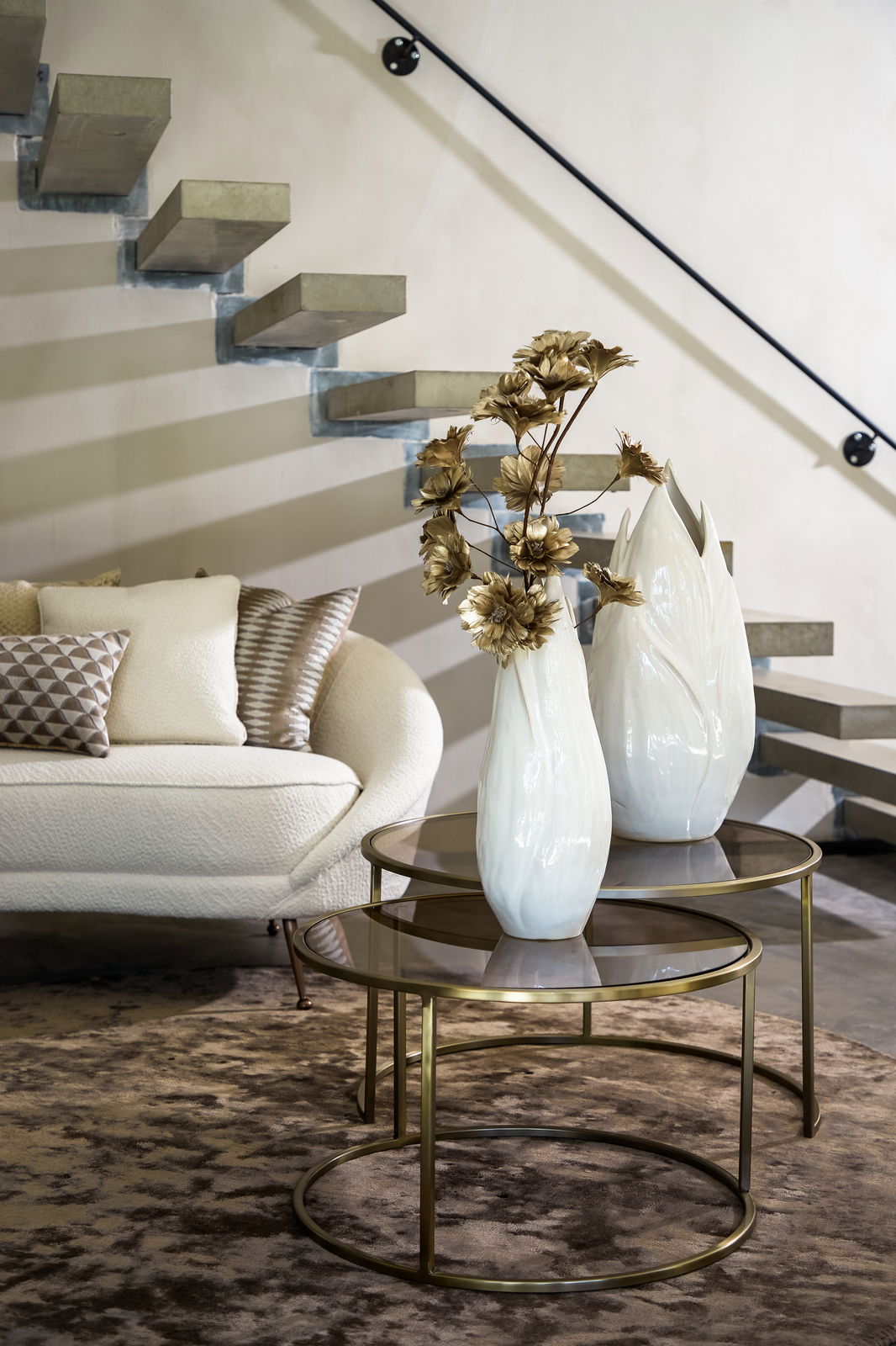 Light&Living Coffee-Table Set SAISON Zwei runde Couchtische mit Glasplatten und goldenen Metallrahmen, dekoriert mit weißen Vasen und goldenen Blumen, in einem modernen Wohnzimmer mit Treppe im Hintergrund, seitliche Perspektive.
