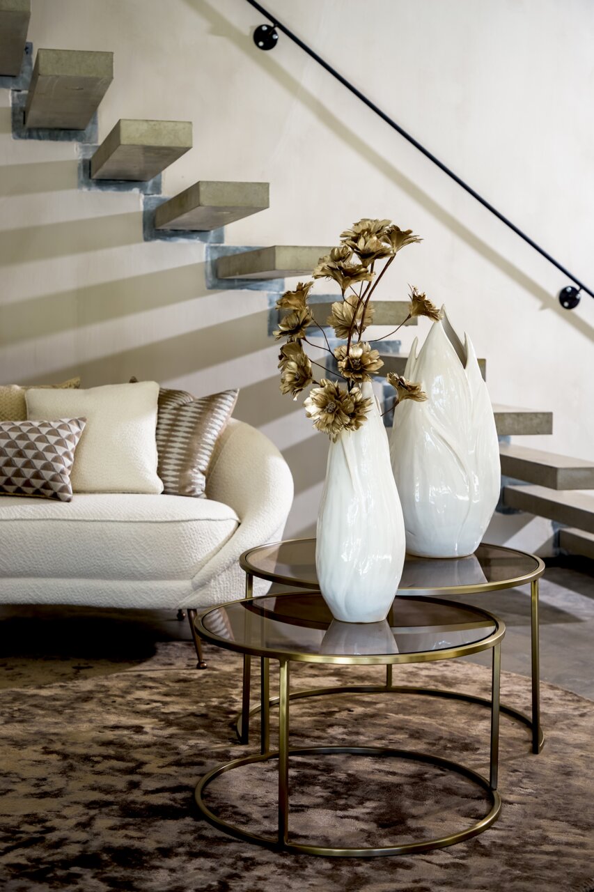 Zwei runde Couchtische mit Glasplatten und goldenen Metallrahmen, dekoriert mit weißen Vasen und goldenen Blumen, in einem modernen Wohnzimmer mit Treppe im Hintergrund, seitliche Perspektive.