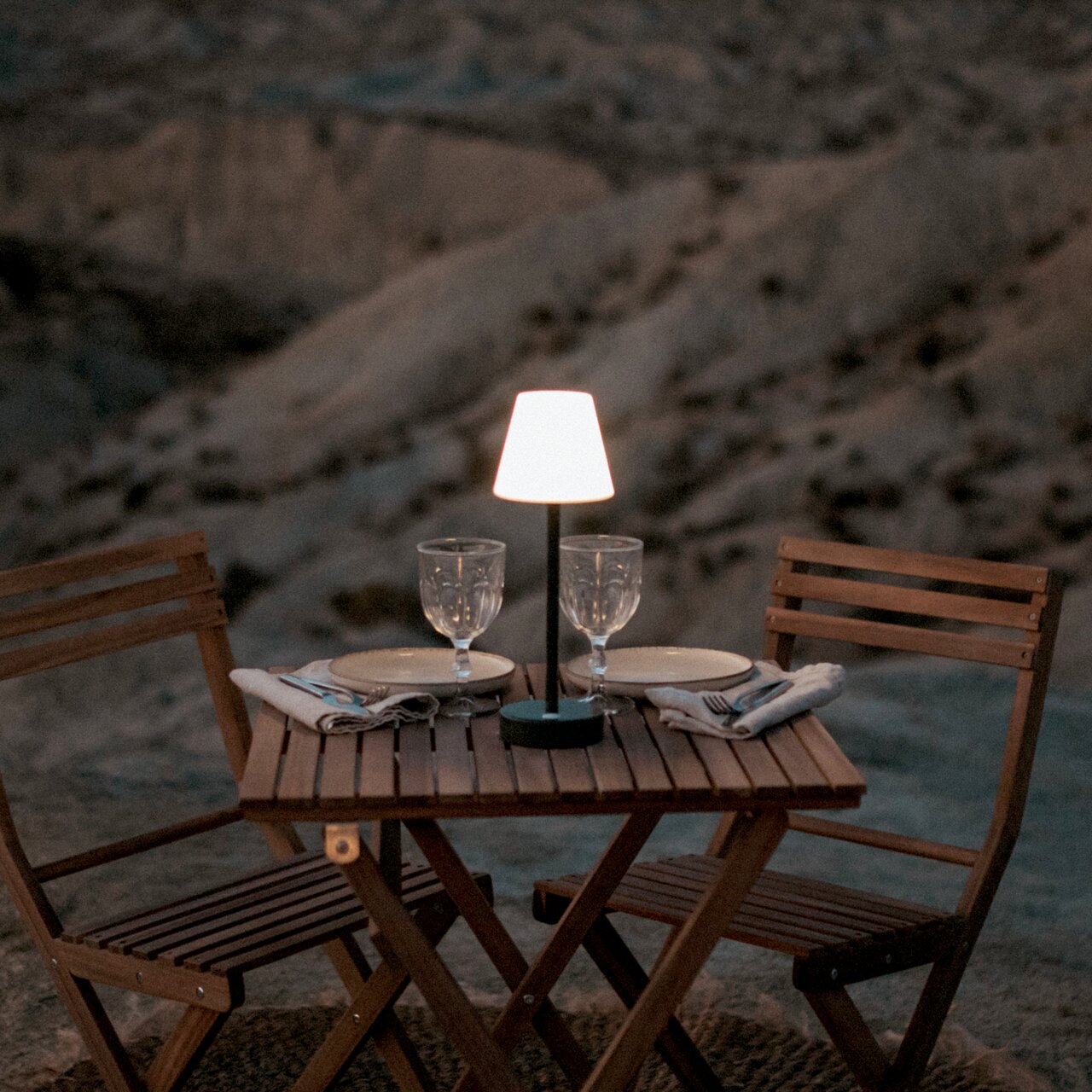 Kleine draadloze LED-tafellamp met witte lampenkap op een gedekte houten tafel buiten tussen twee houten stoelen, van voren gefotografeerd vanaf ooghoogte.