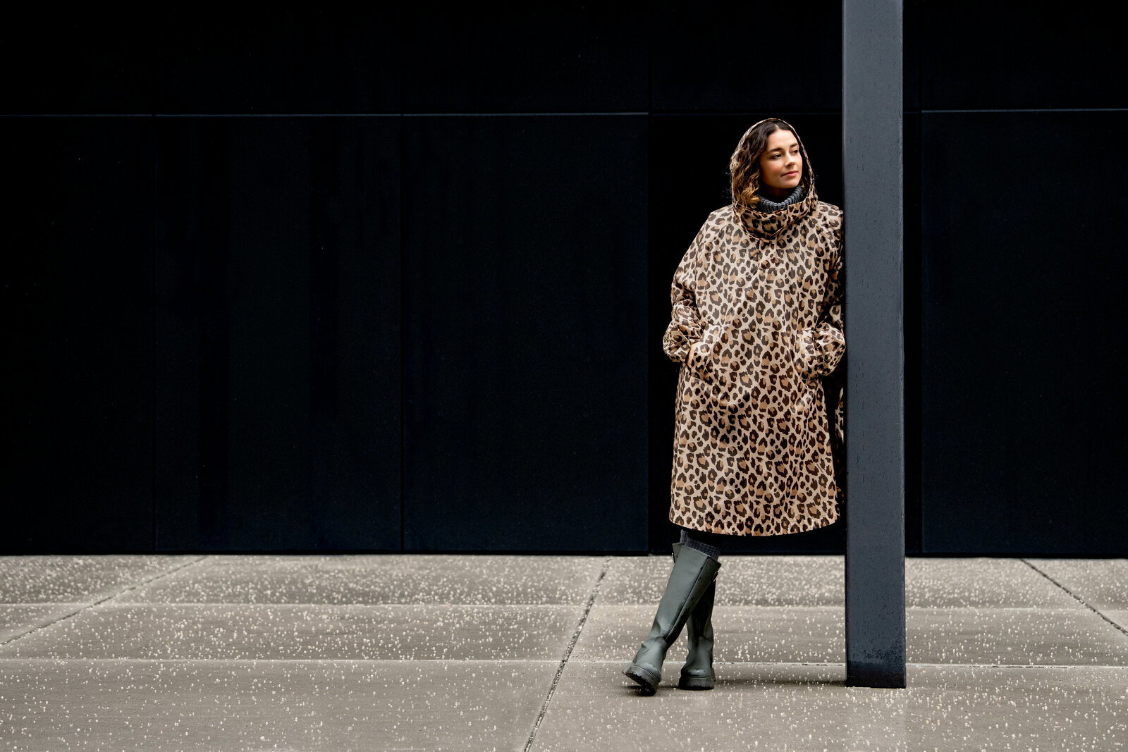 Frau in langem Leopardenmuster-Mantel mit hohem Kragen und Taschen, seitlich an einer dunklen Wand neben einer Säule stehend, aufgenommen in seitlicher Ganzkörperansicht.