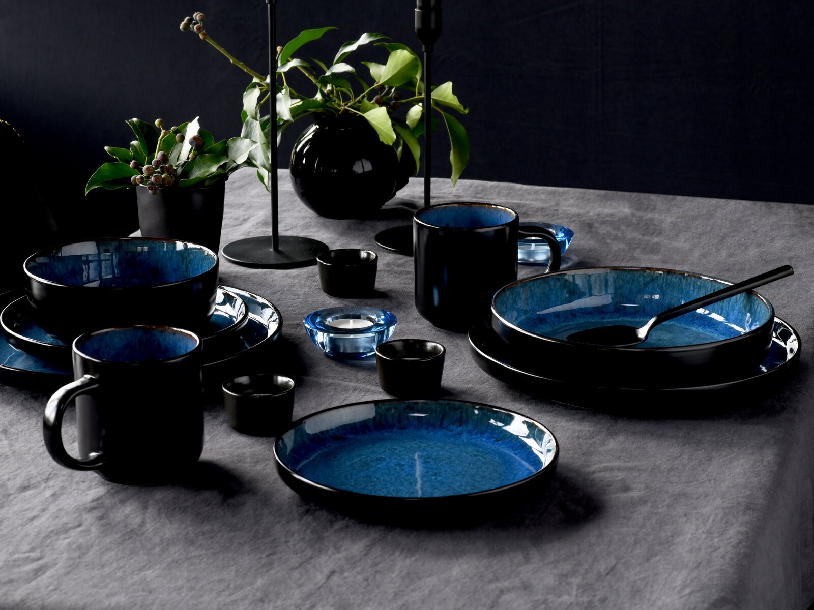 Elegantes Kombiservice 16-tlg in Schwarz und Blau, bestehend aus Tellern, Schalen und Tassen, auf einem gedeckten Tisch mit Pflanzen und Kerzenhaltern, seitliche Perspektive.