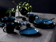 Elegantes Kombiservice 16-tlg in Schwarz und Blau, bestehend aus Tellern, Schalen und Tassen, auf einem gedeckten Tisch mit Pflanzen und Kerzenhaltern, seitliche Perspektive.