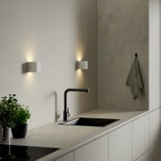 Moderne weiße, würfelförmige Wandleuchte mit Up- und Downlight über einer Küchenarbeitsplatte mit schwarzer Armatur und Spüle, schräger Perspektive von vorne rechts