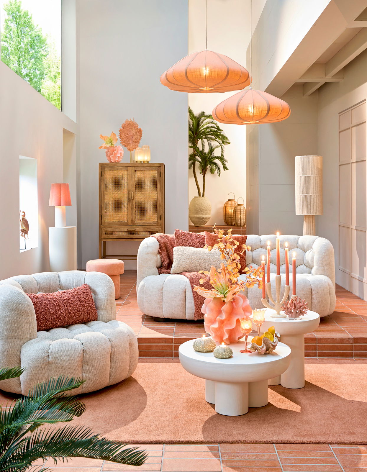 Gemütliche Wohnzimmer-Szene mit bequemen, cremefarbenen Sesseln und Sofas, dekoriert mit orangefarbenen Kissen. Im Vordergrund stehen runde, weiße Couchtische mit dekorativen Elementen. Die Perspektive zeigt den Raum von vorne, mit einem Blick auf die ges