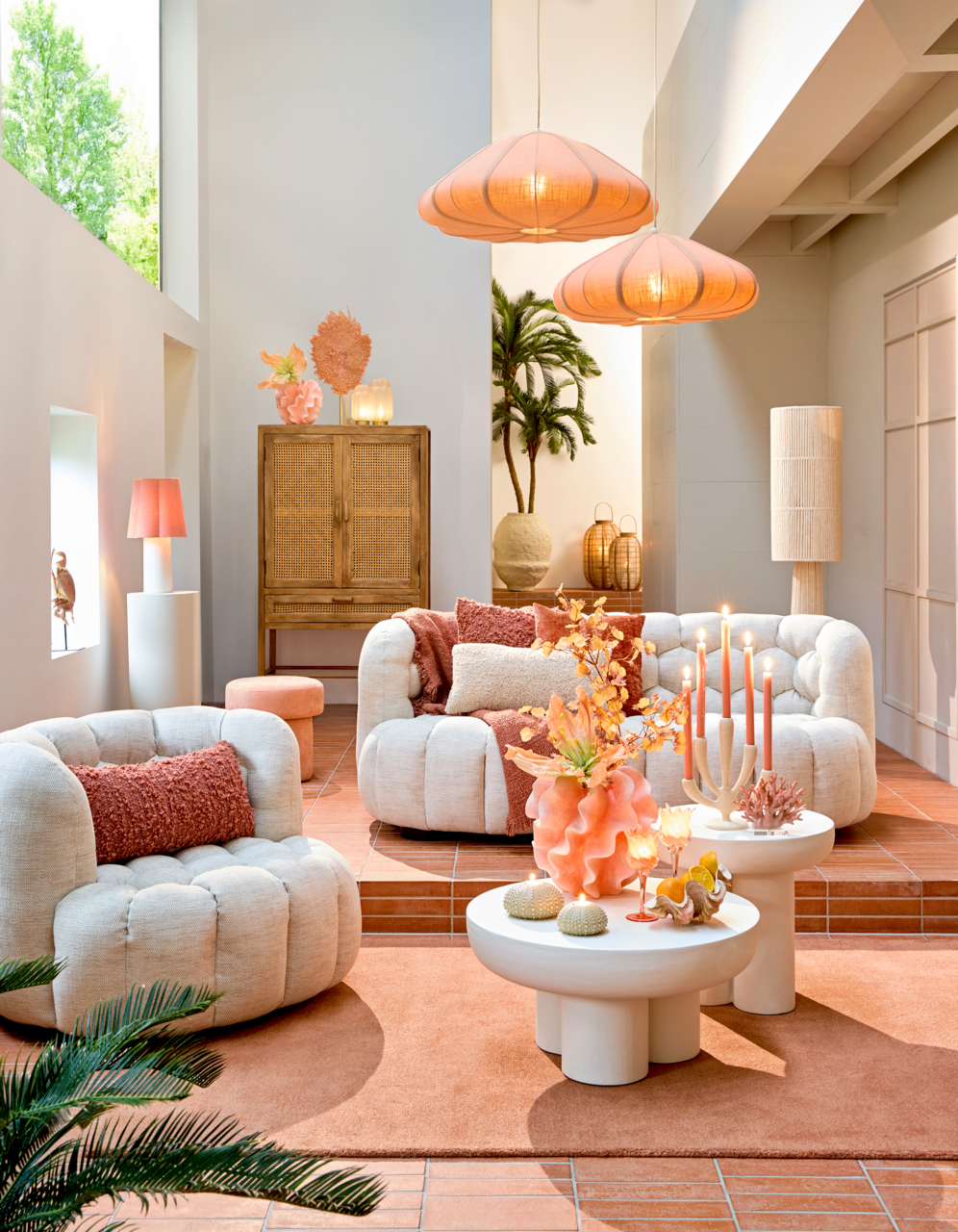 Gemütliche Wohnzimmer-Szene mit bequemen, cremefarbenen Sesseln und Sofas, dekoriert mit orangefarbenen Kissen. Im Vordergrund stehen runde, weiße Couchtische mit dekorativen Elementen. Die Perspektive zeigt den Raum von vorne, mit einem Blick auf die ges