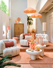 Gemütliche Wohnzimmer-Szene mit bequemen, cremefarbenen Sesseln und Sofas, dekoriert mit orangefarbenen Kissen. Im Vordergrund stehen runde, weiße Couchtische mit dekorativen Elementen. Die Perspektive zeigt den Raum von vorne, mit einem Blick auf die ges