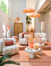 Gemütliche Wohnzimmer-Szene mit bequemen, cremefarbenen Sesseln und Sofas, dekoriert mit orangefarbenen Kissen. Im Vordergrund stehen runde, weiße Couchtische mit dekorativen Elementen. Die Perspektive zeigt den Raum von vorne, mit einem Blick auf die ges