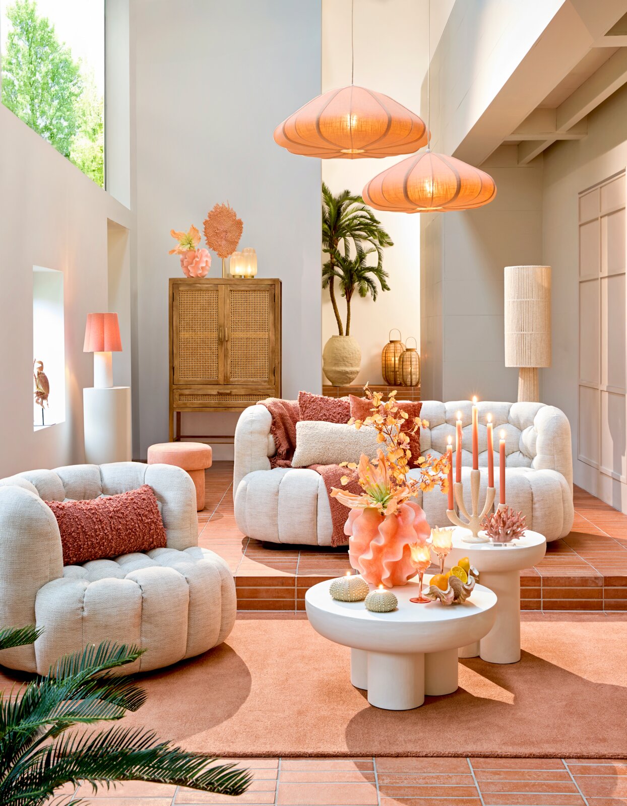 Gemütliche Wohnzimmer-Szene mit bequemen, cremefarbenen Sesseln und Sofas, dekoriert mit orangefarbenen Kissen. Im Vordergrund stehen runde, weiße Couchtische mit dekorativen Elementen. Die Perspektive zeigt den Raum von vorne, mit einem Blick auf die ges