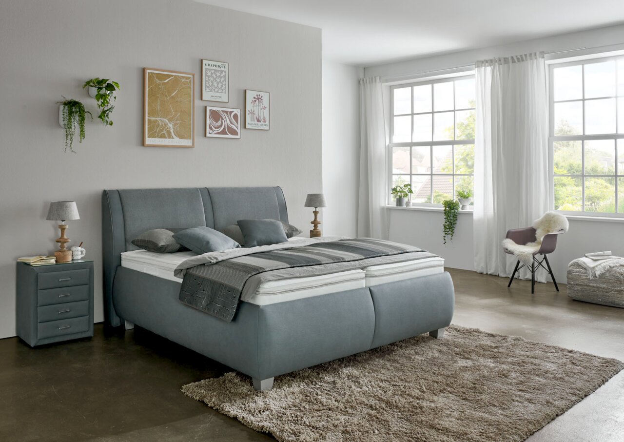 Modernes Polsterbett in einem hellen Schlafzimmer, seitliche Perspektive, mit grauem Kopfteil und passendem Nachttisch, dekoriert mit Kissen und Decken.