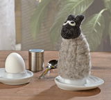 Eierwärmer in Form eines Schafs aus Filz, frontal auf einem Tisch neben einem Ei und einem Löffel platziert.