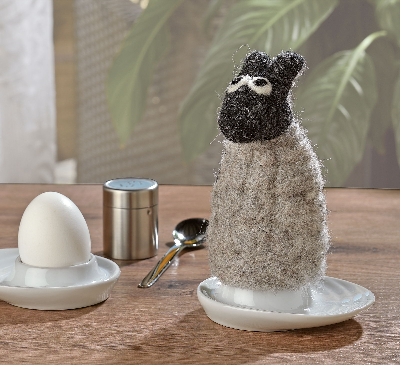 Eierwärmer in Form eines Schafs aus Filz, frontal auf einem Tisch neben einem Ei und einem Löffel platziert.