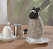 Eierwärmer in Form eines Schafs aus Filz, frontal auf einem Tisch neben einem Ei und einem Löffel platziert.