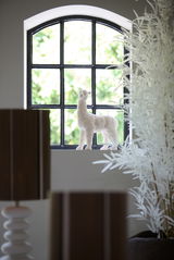 Dekorative weiße Lama-Figur auf einer Fensterbank, von der Seite betrachtet, mit einem Fenster im Hintergrund.