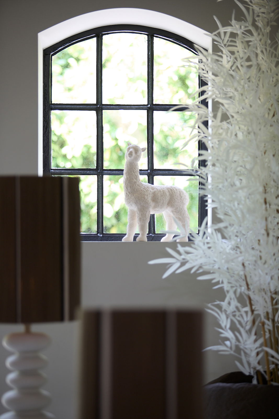 Dekorative weiße Lama-Figur auf einer Fensterbank, von der Seite betrachtet, mit einem Fenster im Hintergrund.