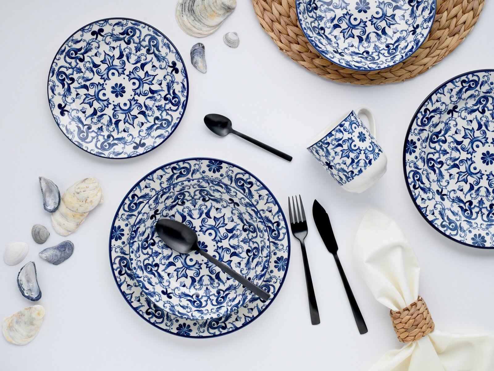 Flachlay aus der Vogelperspektive: blau-weiß gemustertes Keramikgeschirr (Teller und Schale) mit passender Tasse, schwarzem Besteck, Muscheln und geflochtenen Untersetzern auf weißem Hintergrund
